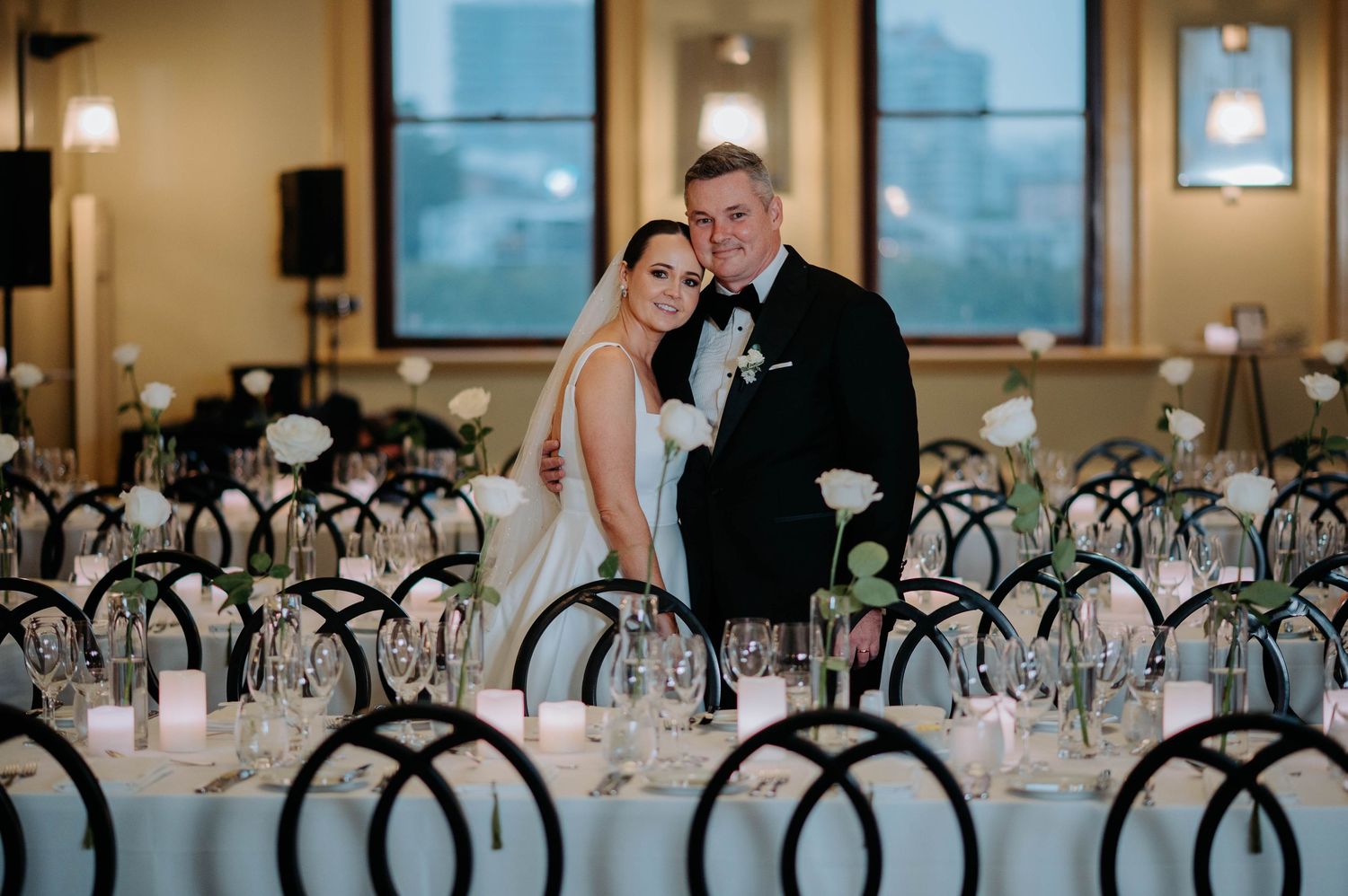 A romantic couple poses in formal wedding attire inside an elegant reception hall with black bentwood chairs and candlelit tables.
