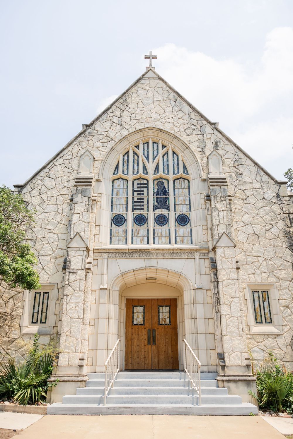 Stone church entrance with gothic-style arched doorway and decorative window features a cross on top of its facade.