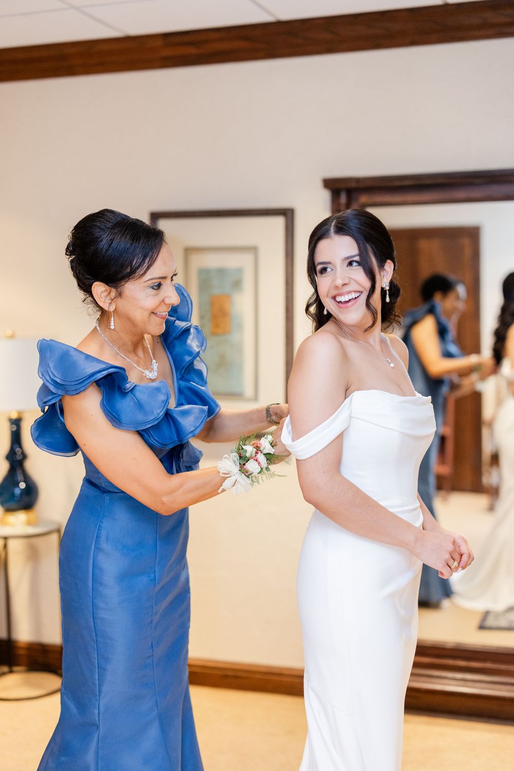 A woman in a royal blue dress helps adjust details on a bride's elegant white wedding gown.