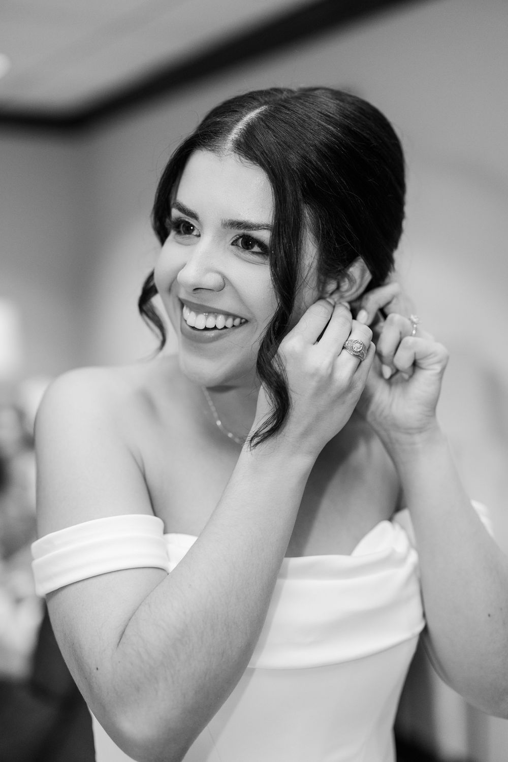 A bride in an off-shoulder white wedding dress puts on earrings while smiling in a black and white portrait.