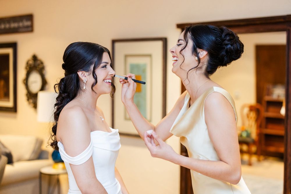 Two women in white dresses laugh together while getting ready for a wedding celebration.
