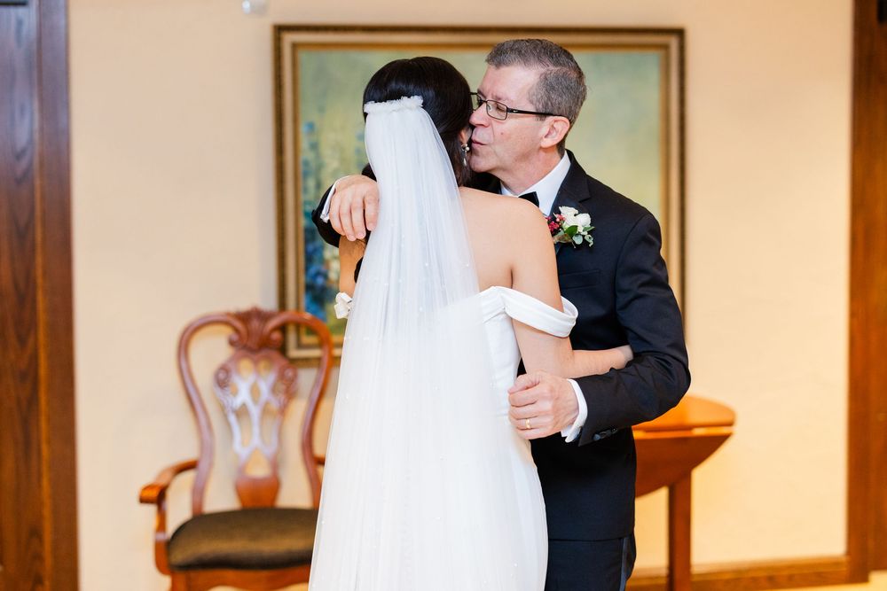 A bride in a white strapless wedding gown embraces someone wearing a black tuxedo in an elegant indoor setting.