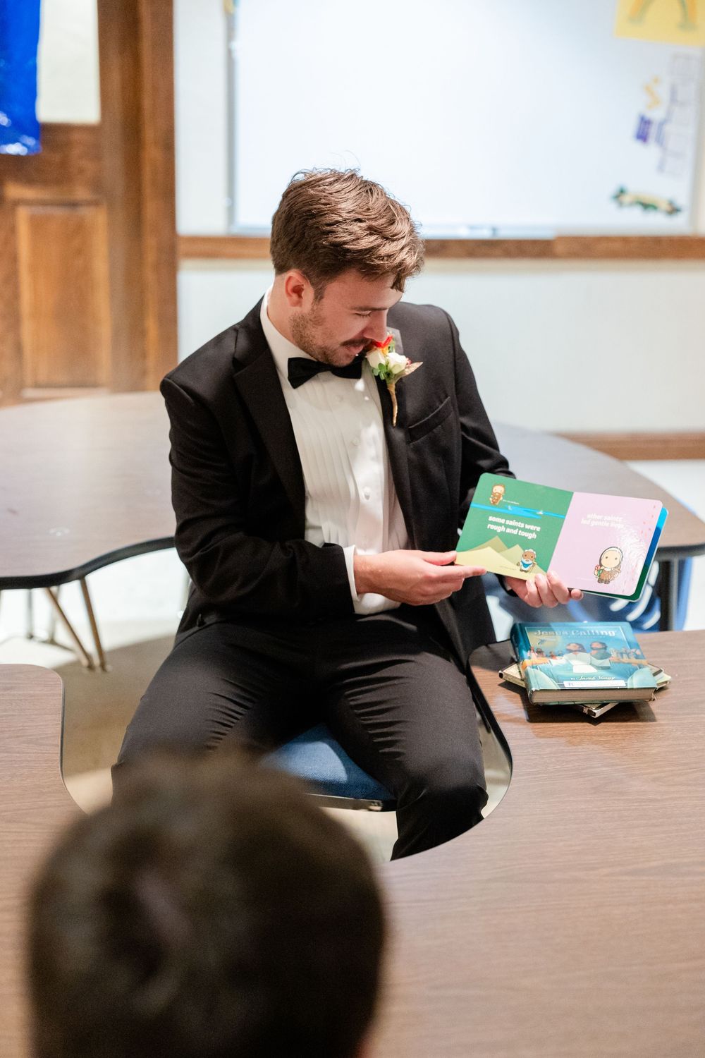 A person in a tuxedo sits in a classroom reading a children's book during a special event.