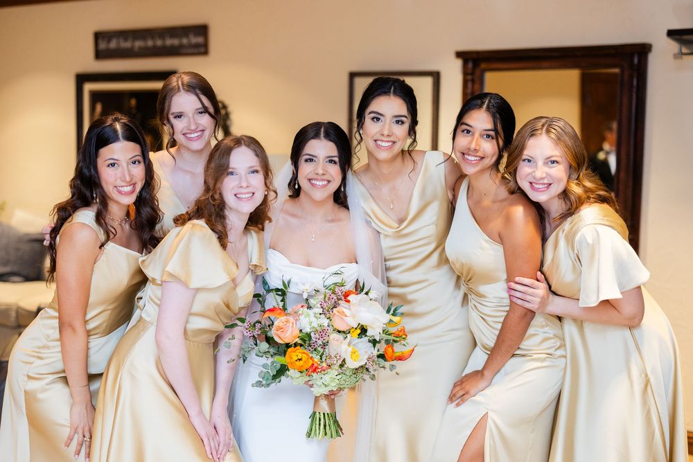 Bridesmaids in champagne dresses gather around bride holding vibrant orange and white bouquet.