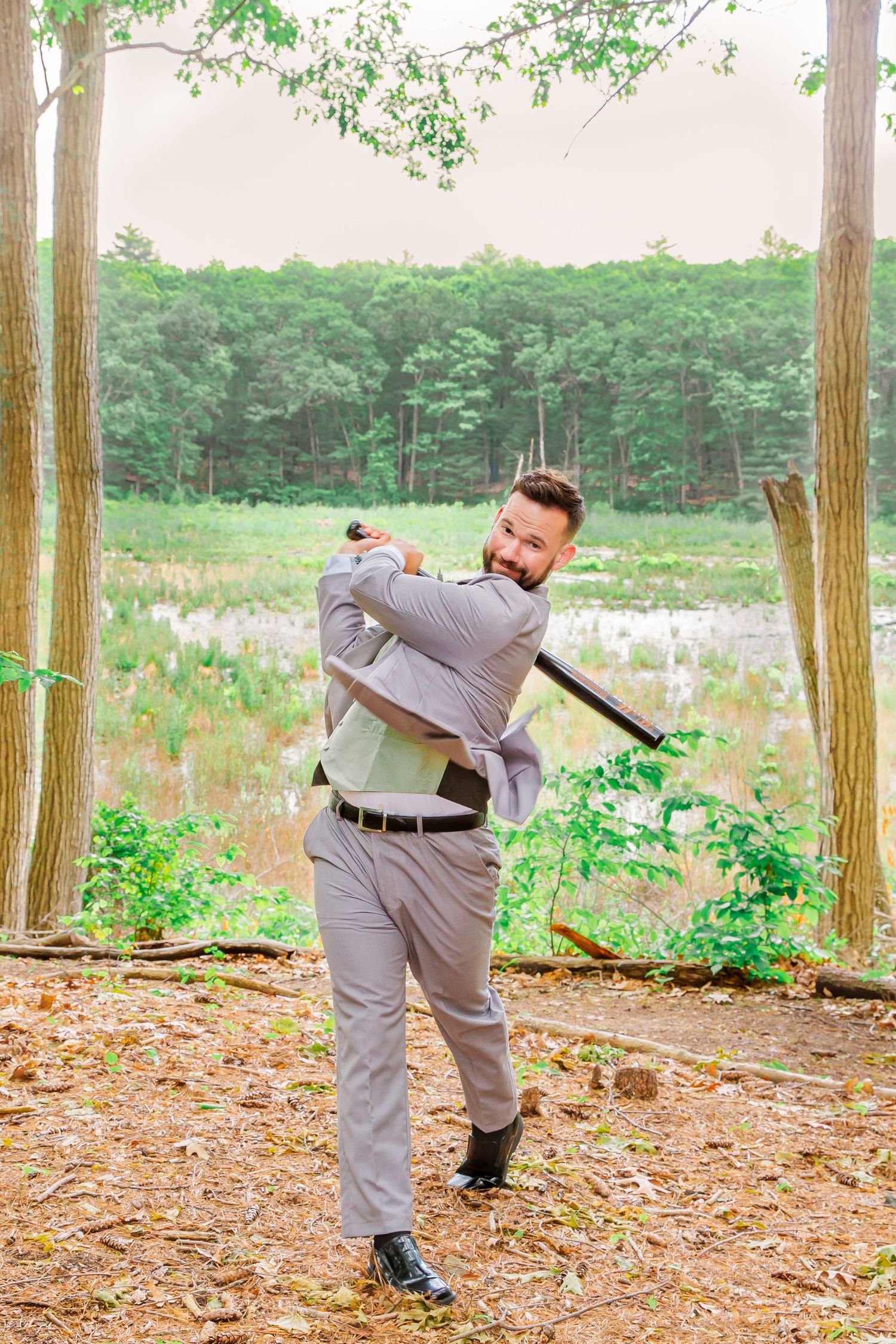 Someone in a gray suit poses with a baseball bat in a wooded area with wildflowers in the background.