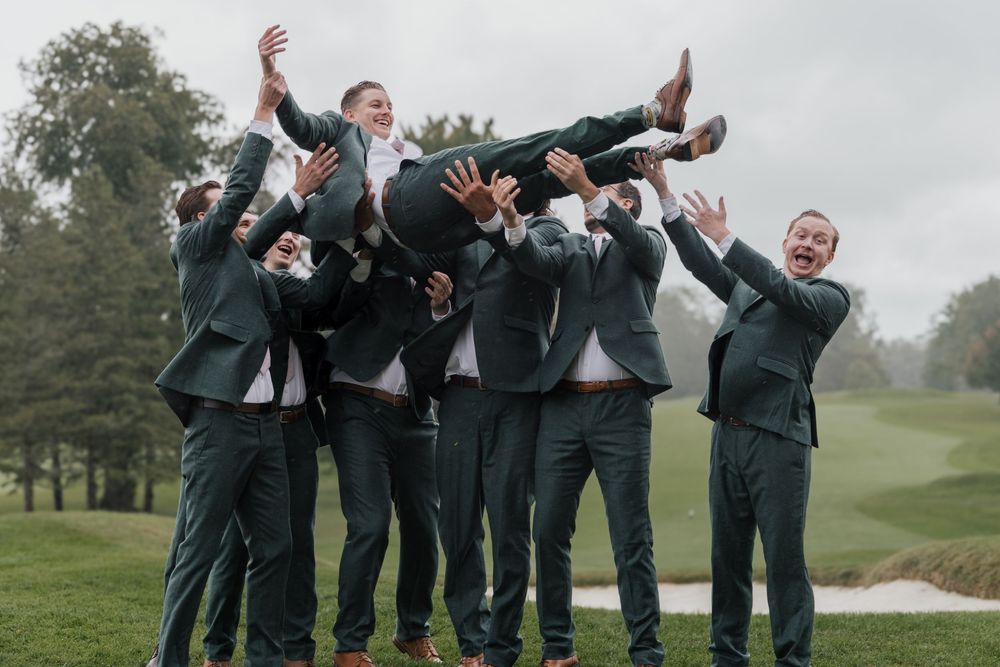 Groomsmen in dark green suits lifting the groom overhead in a playful celebration outdoors.