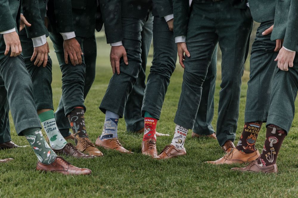 Groomsmen showing off colorful novelty socks with brown dress shoes on green grass.