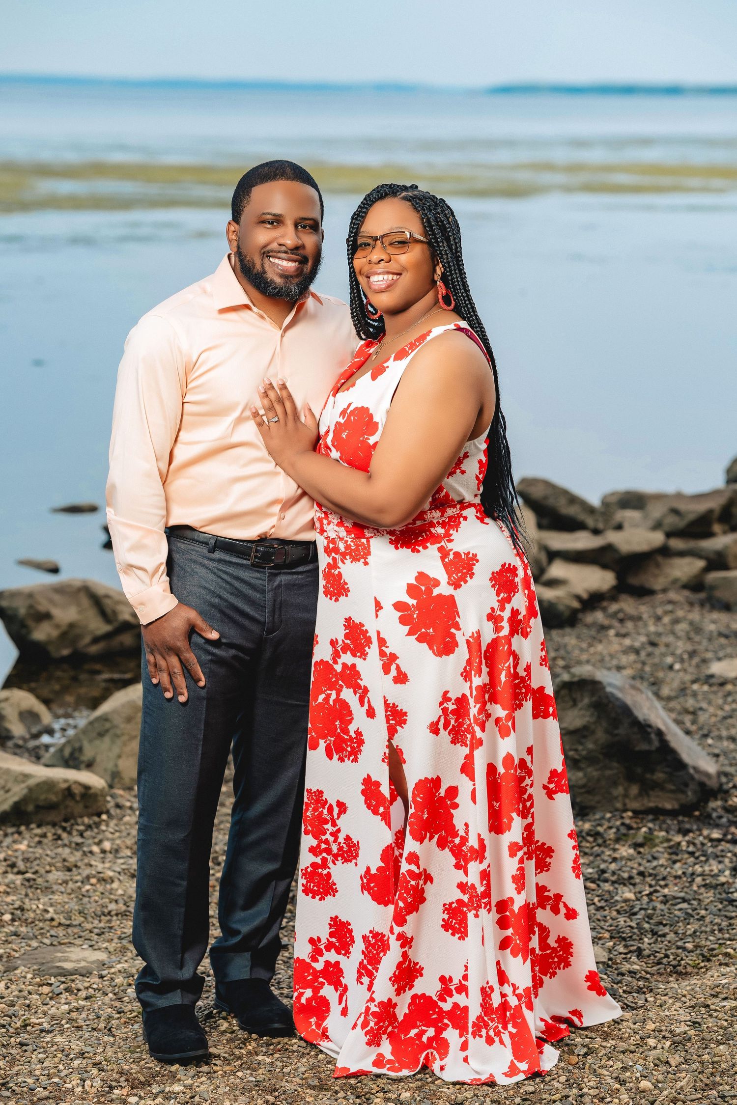 A couple poses together on a rocky beach shore wearing coordinated outfits - a peach shirt and floral red dress.