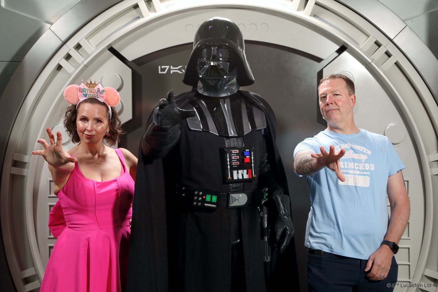 Three people pose in a Star Wars themed photo setting with Darth Vader's costume in the middle of a circular space corridor.