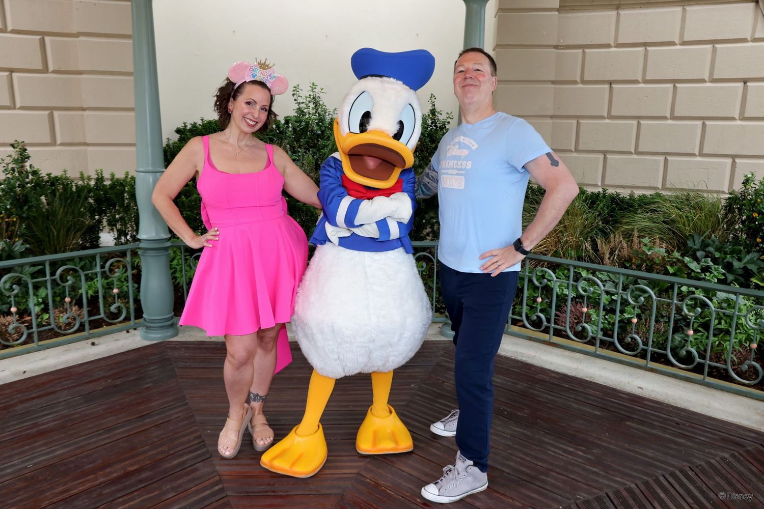 Two people posing with Donald Duck character in sailor outfit at Disney theme park entrance.