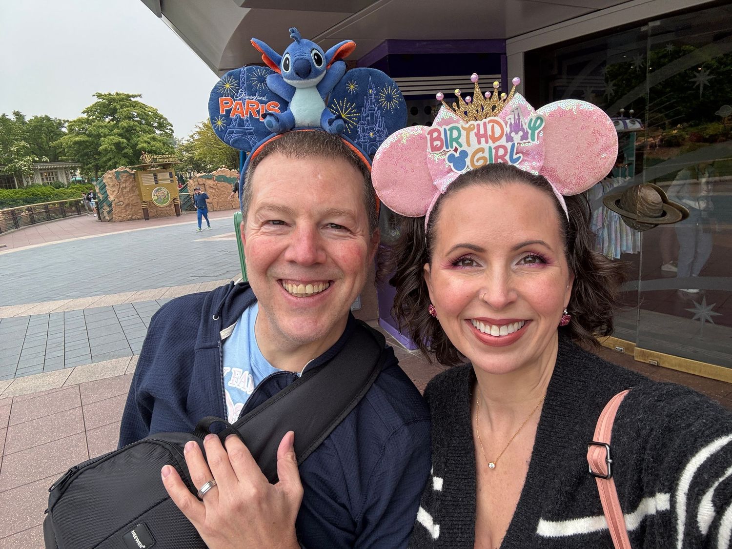 Two Disney park visitors wearing Mickey ears pose together outside, with Stitch-themed and pink sparkly mouse ear headbands.