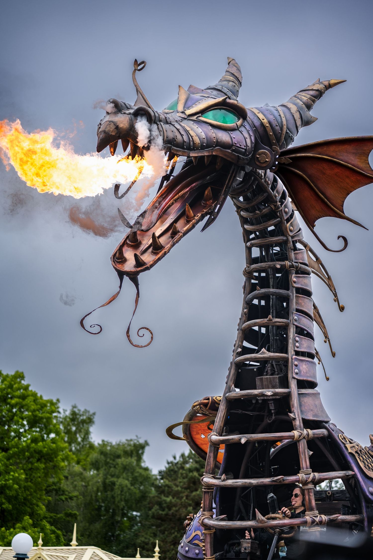 A metallic dragon sculpture with glowing eyes breathes real flames against a cloudy sky at an outdoor event.