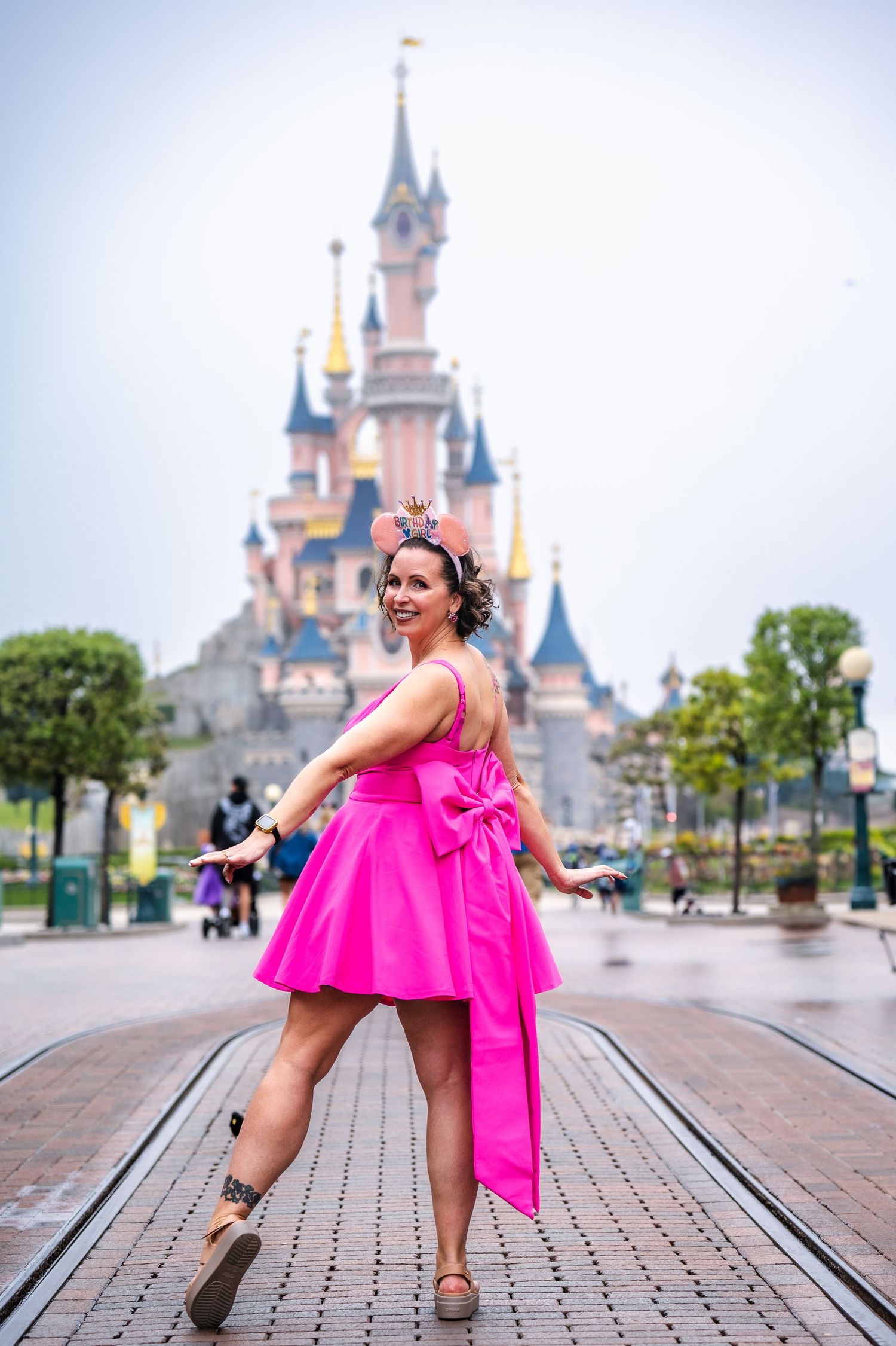 Person in bright pink dress poses cheerfully in front of Disneyland's iconic Sleeping Beauty Castle on a cloudy day.