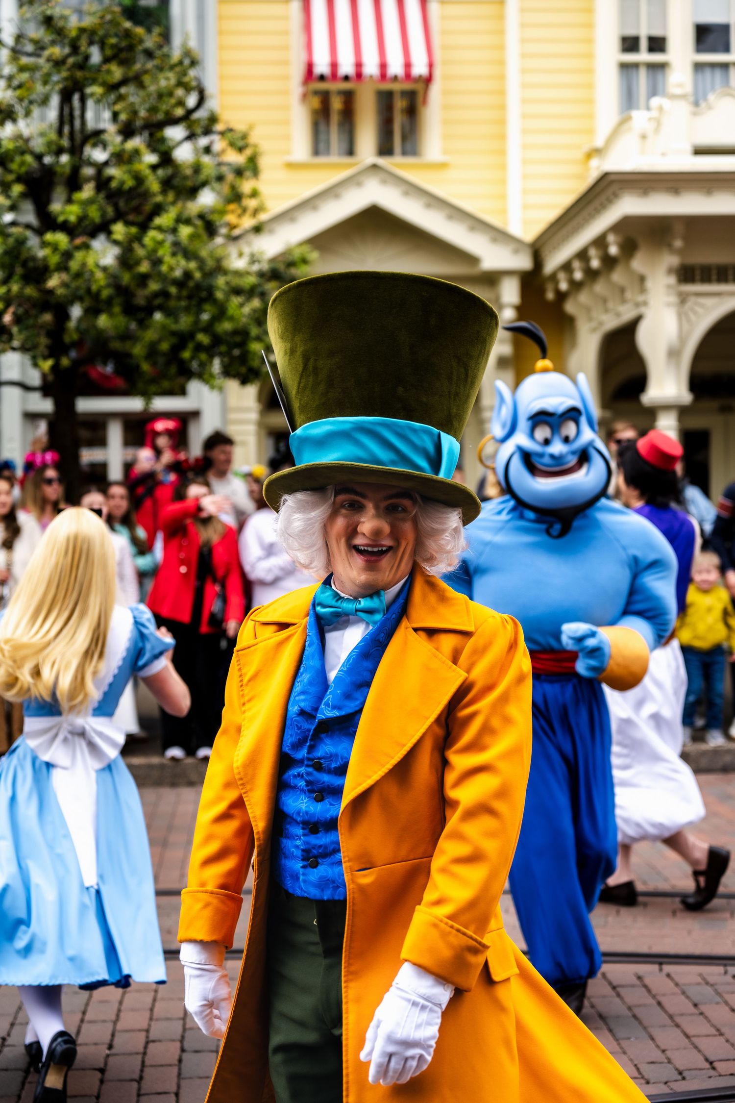 A colorful character in a top hat and bright yellow coat greets visitors at Disneyland with a blue costumed figure behind.