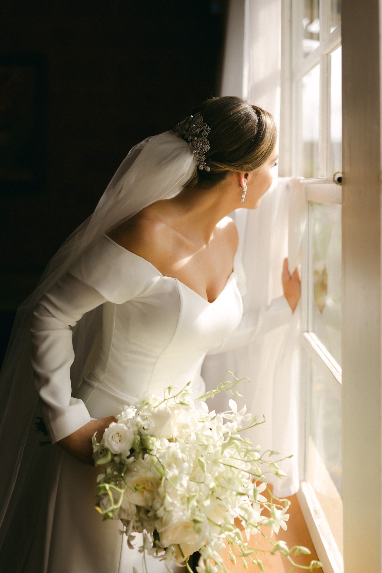 Elegant bride in long-sleeved dress holding white bouquet by window.