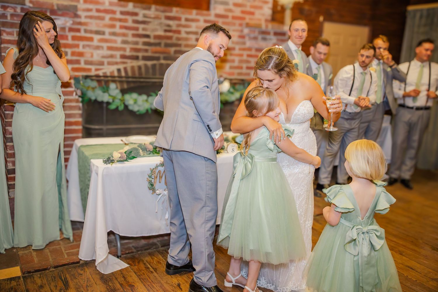 Sweet wedding reception moment with wedding party gathered around cake table in rustic brick-walled venue.