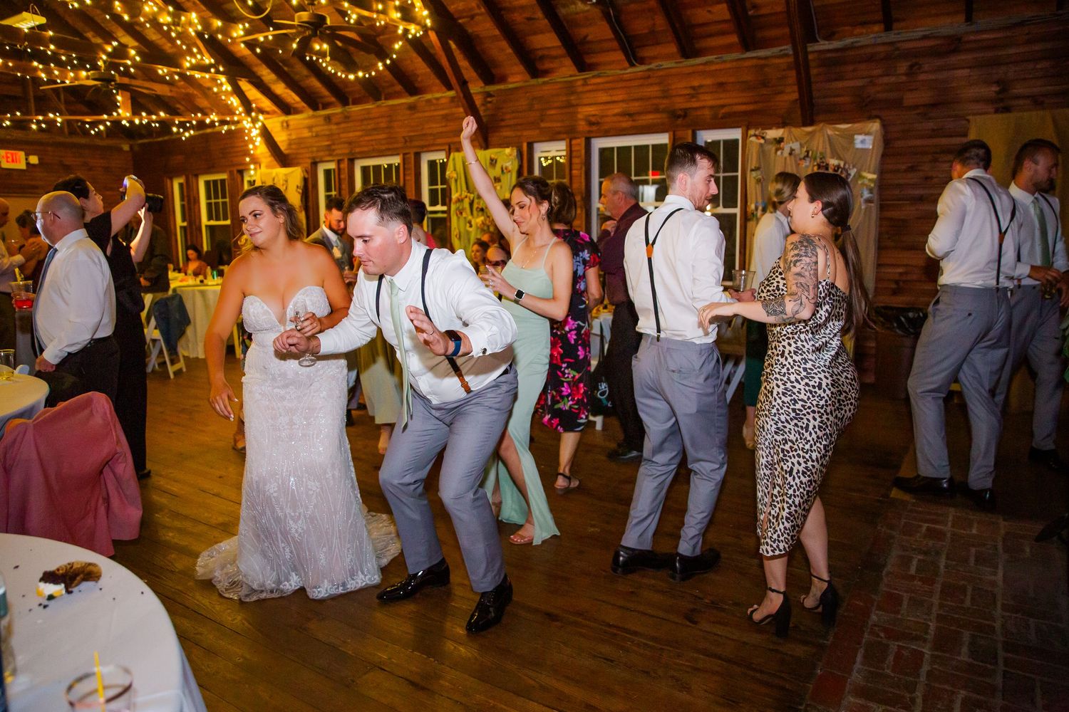 Wedding guests dancing and celebrating on a rustic barn dance floor decorated with string lights at an evening reception.