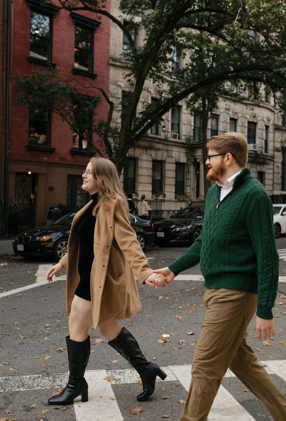 A couple walks playfully through a tree-lined city street on an autumn day wearing coordinated casual outfits.