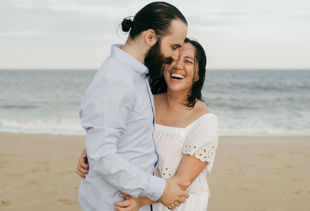 A couple embraces and laughs together during a romantic beach photoshoot wearing white outfits.