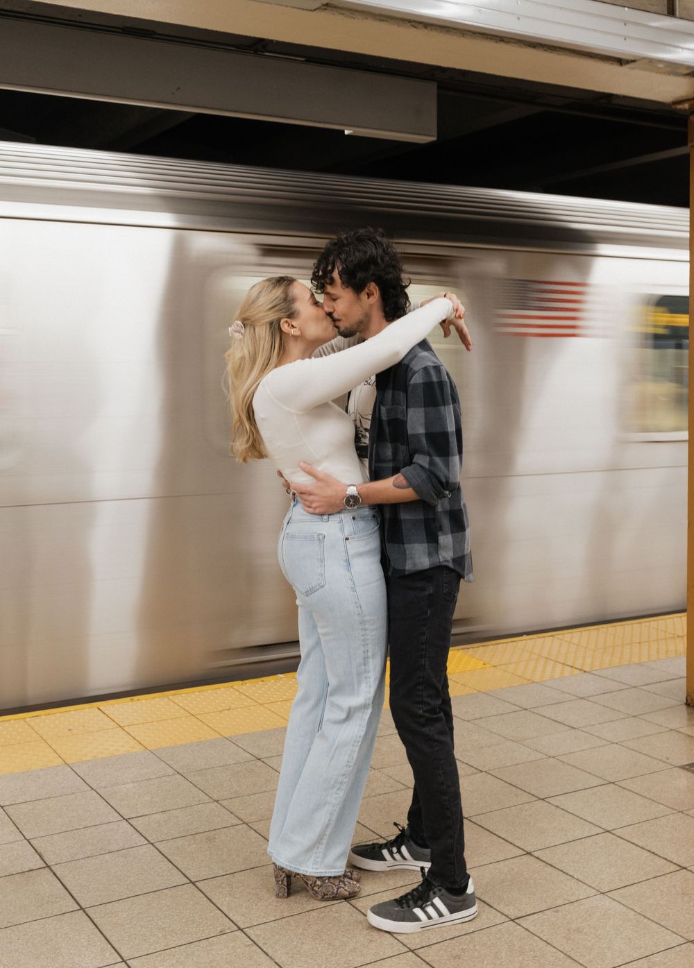 A couple embraces and dances together on a subway platform as a train blurs past in the background.