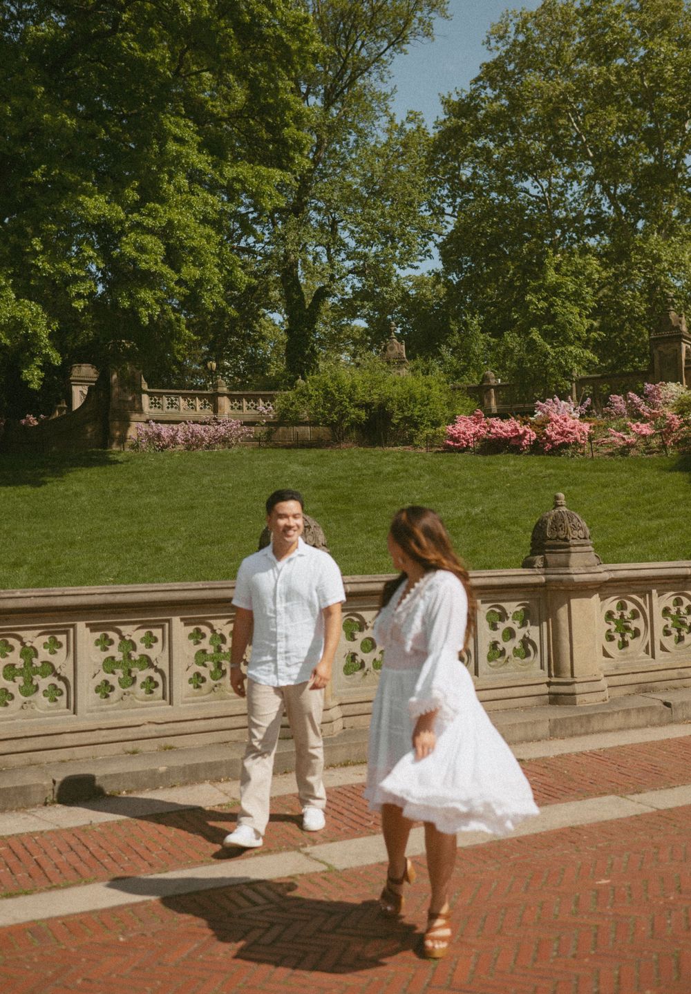 A couple in white outfits walking on brick steps near a stone balustrade with blooming azaleas and trees in the background.