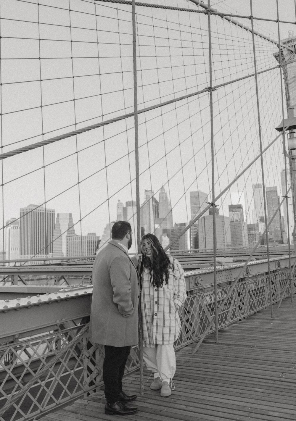 A couple walks along the Brooklyn Bridge walkway with the New York City skyline visible in the background.