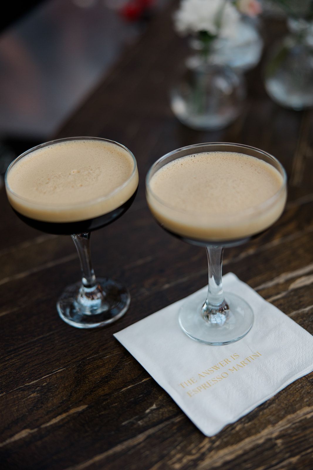 Two creamy espresso martinis served in coupe glasses on a dark wooden table with a white cocktail napkin.