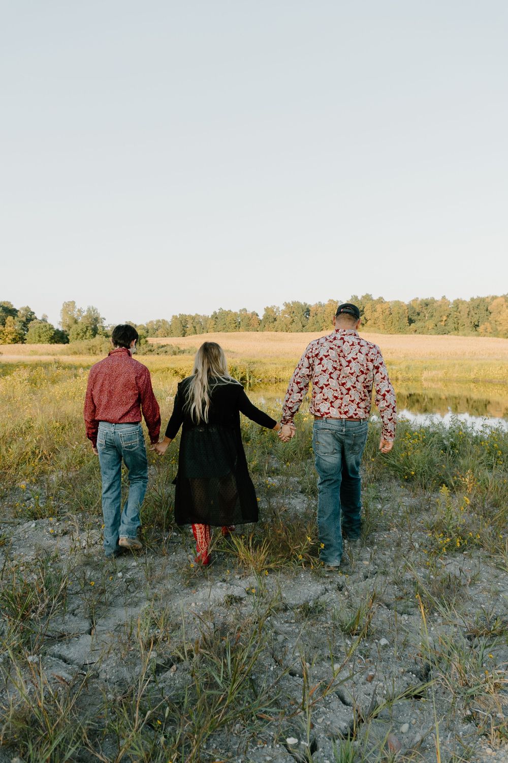 Three people walk away from camera through a grassy field at sunset in a casual outdoor setting.