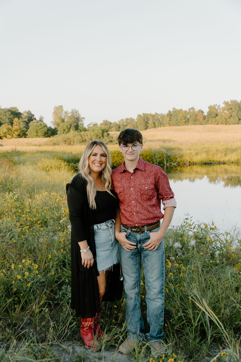 A couple stands together in a field of wildflowers next to a serene pond during golden hour.