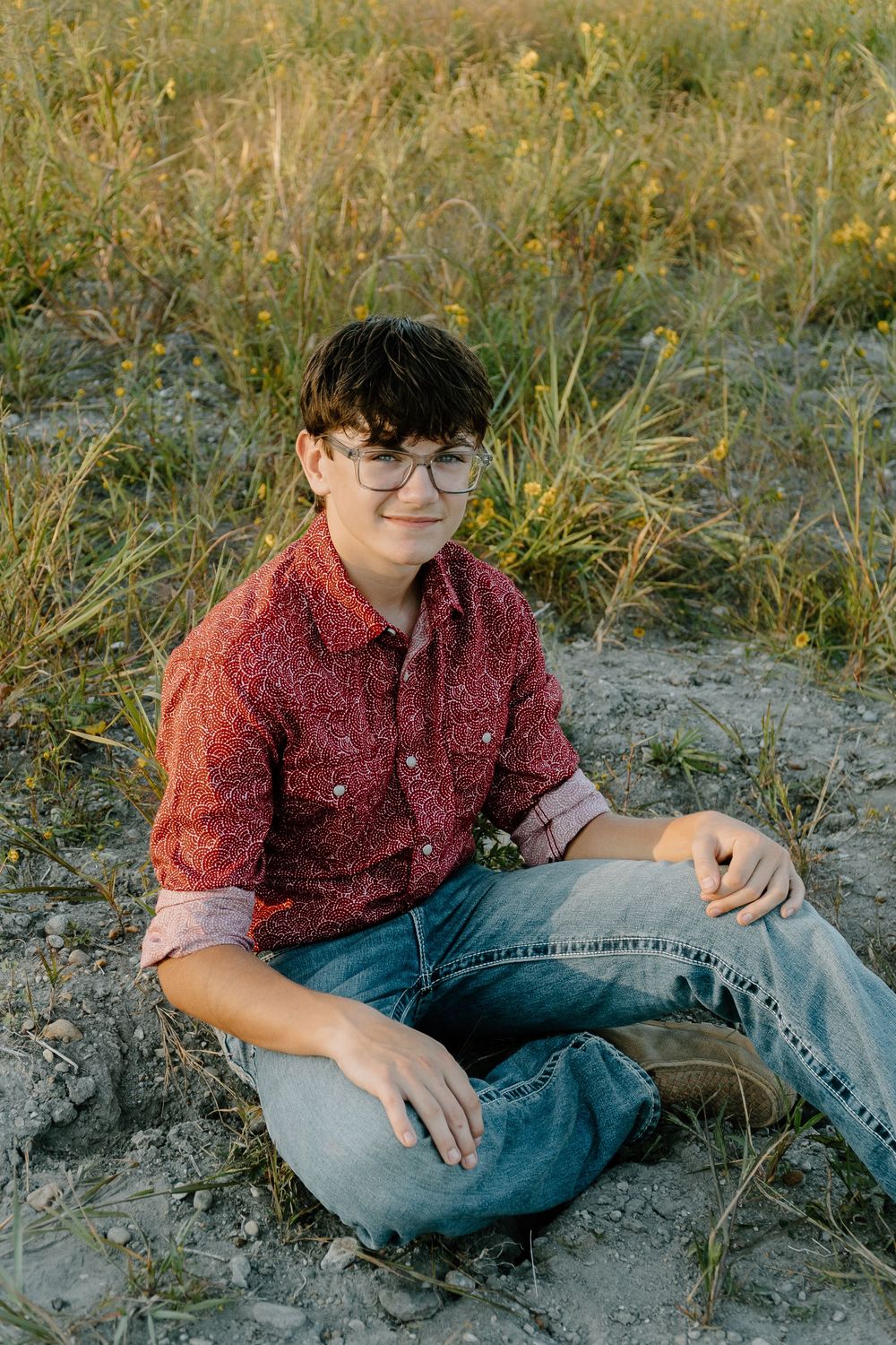 A person in a burgundy button-down shirt and jeans sits casually in tall grass during golden hour.
