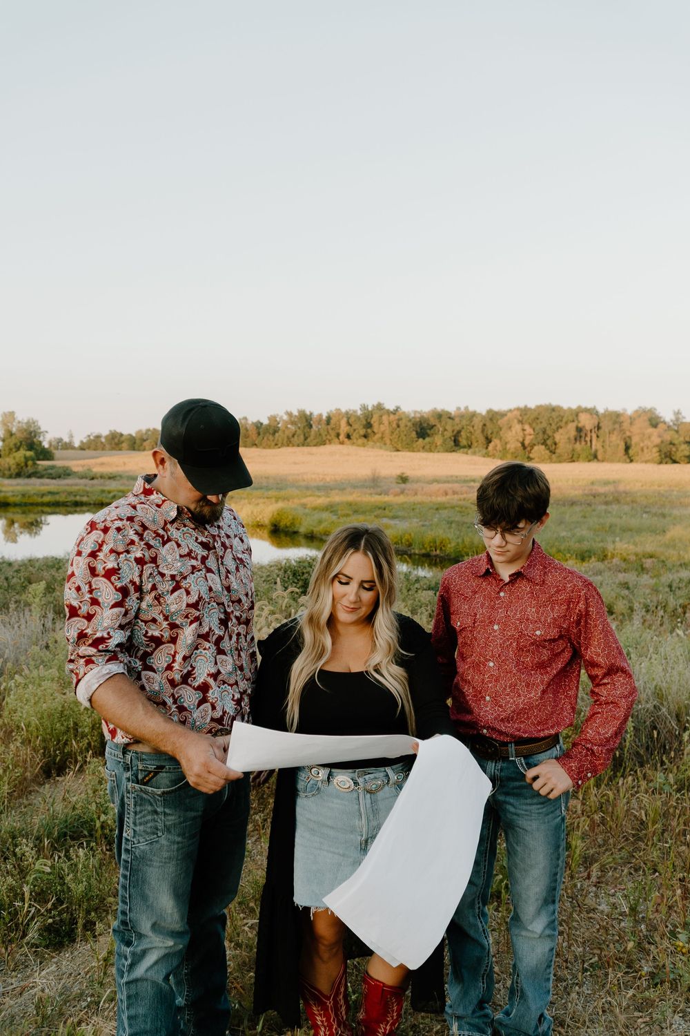 A couple in casual western attire playfully poses in a golden field at sunset with a map.