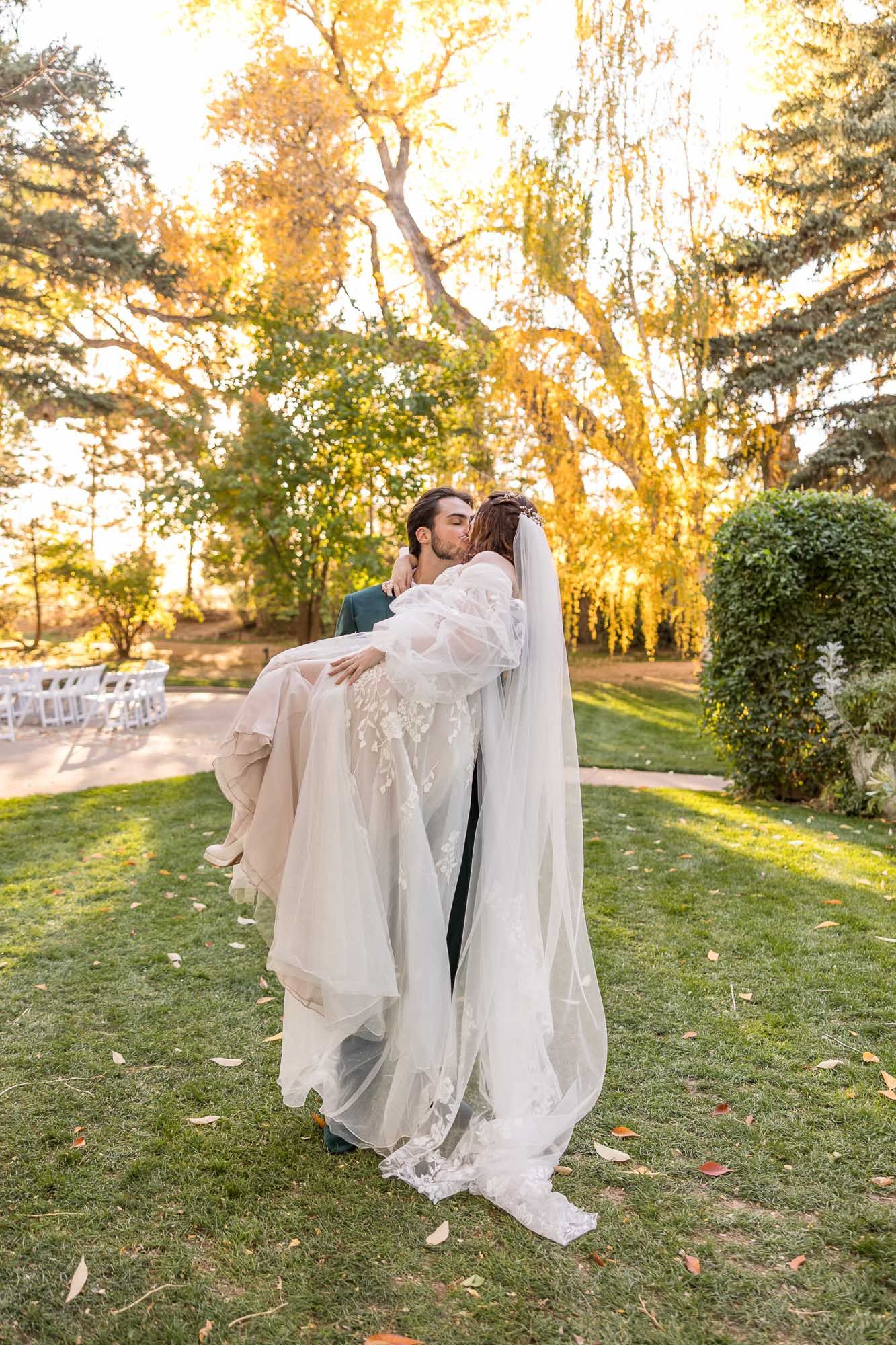 A romantic wedding couple embraces in a garden setting with golden autumn trees and sunlight glowing in the background.