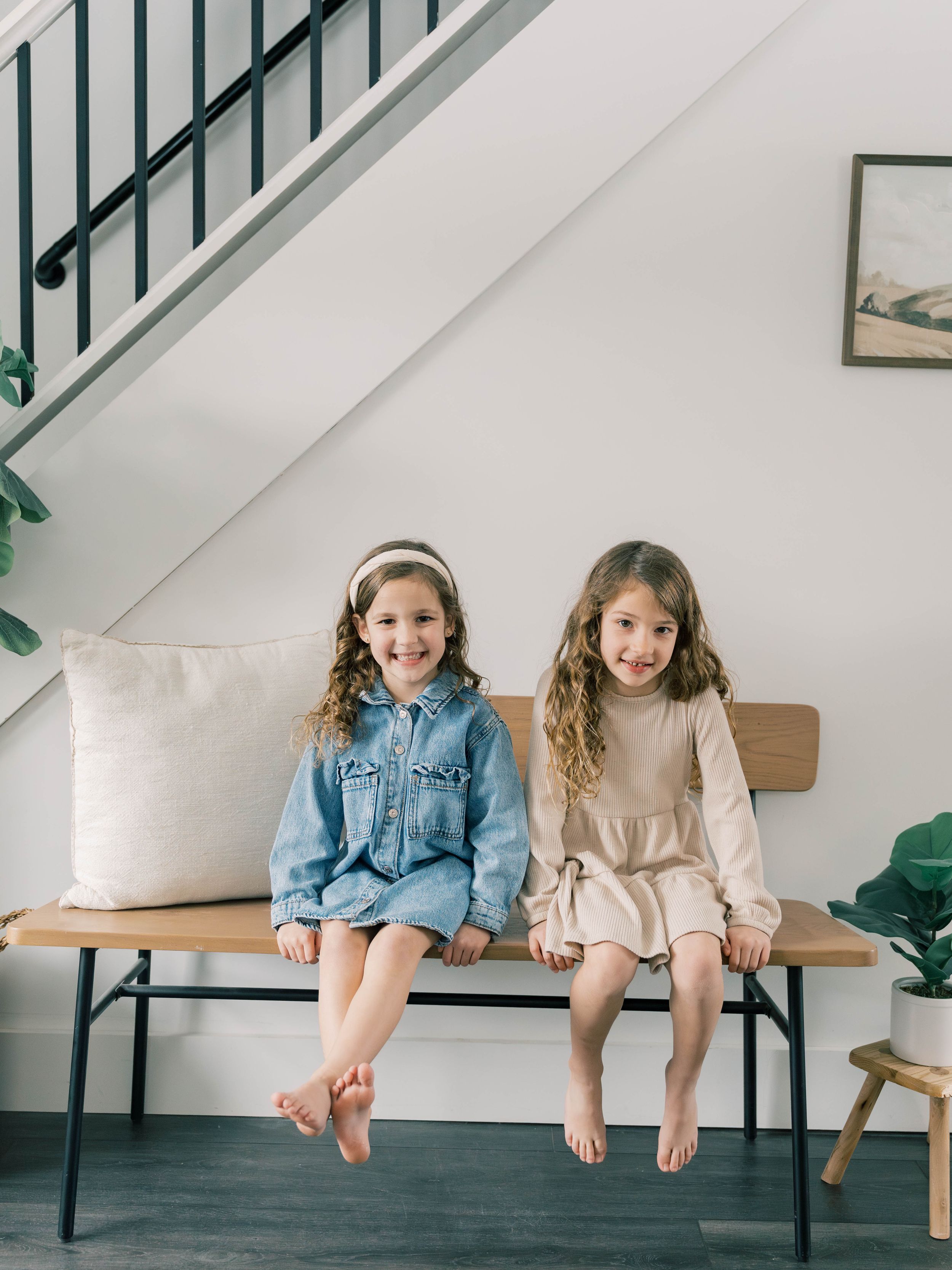 Two children sitting together on a modern bench with white cushions under a staircase in a minimalist home interior.