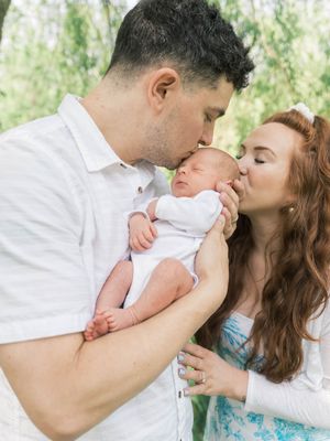 A couple in white and turquoise outfits share tender moments with their newborn baby in a bright outdoor setting.