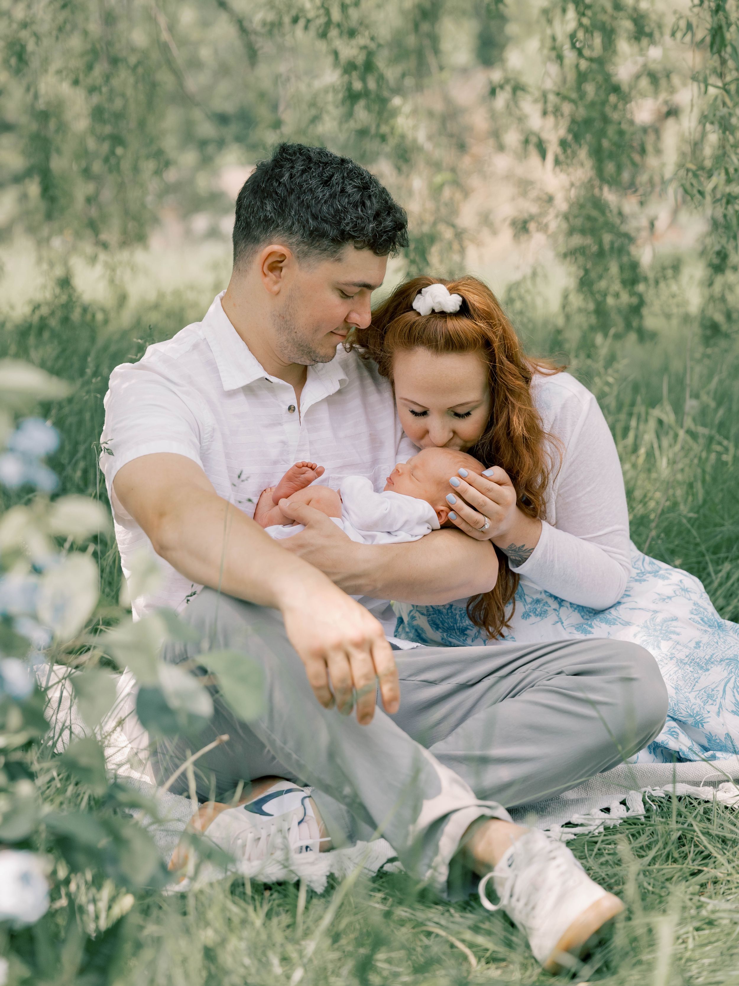 A couple sits in a meadow on a sunny day, tenderly cradling a newborn baby wrapped in white blankets.