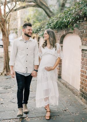 Expectant couple in light colored outfits walking together on a brick path with arches and greenery in the background.