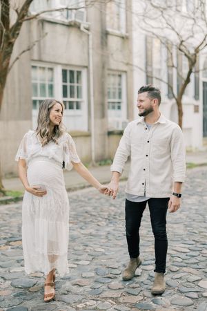 A couple in white outfits walks playfully together on a cobblestone path lined with trees on a cloudy day.