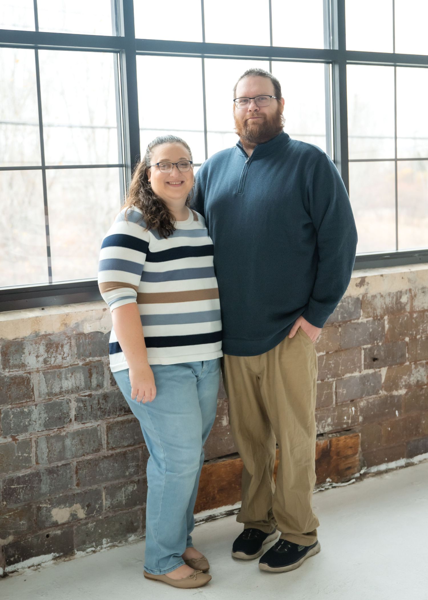 A couple stands together in a bright industrial space with exposed brick walls wearing matching striped shirts.