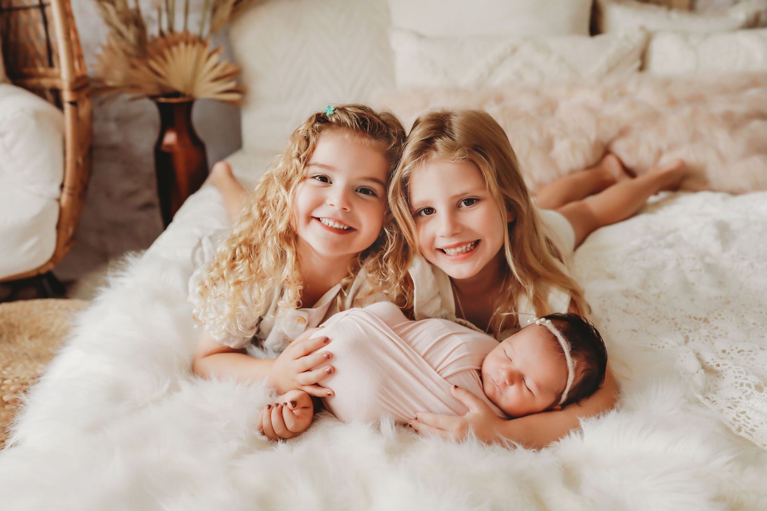Three siblings share a tender moment lying together on a cozy white blanket in warm lighting.