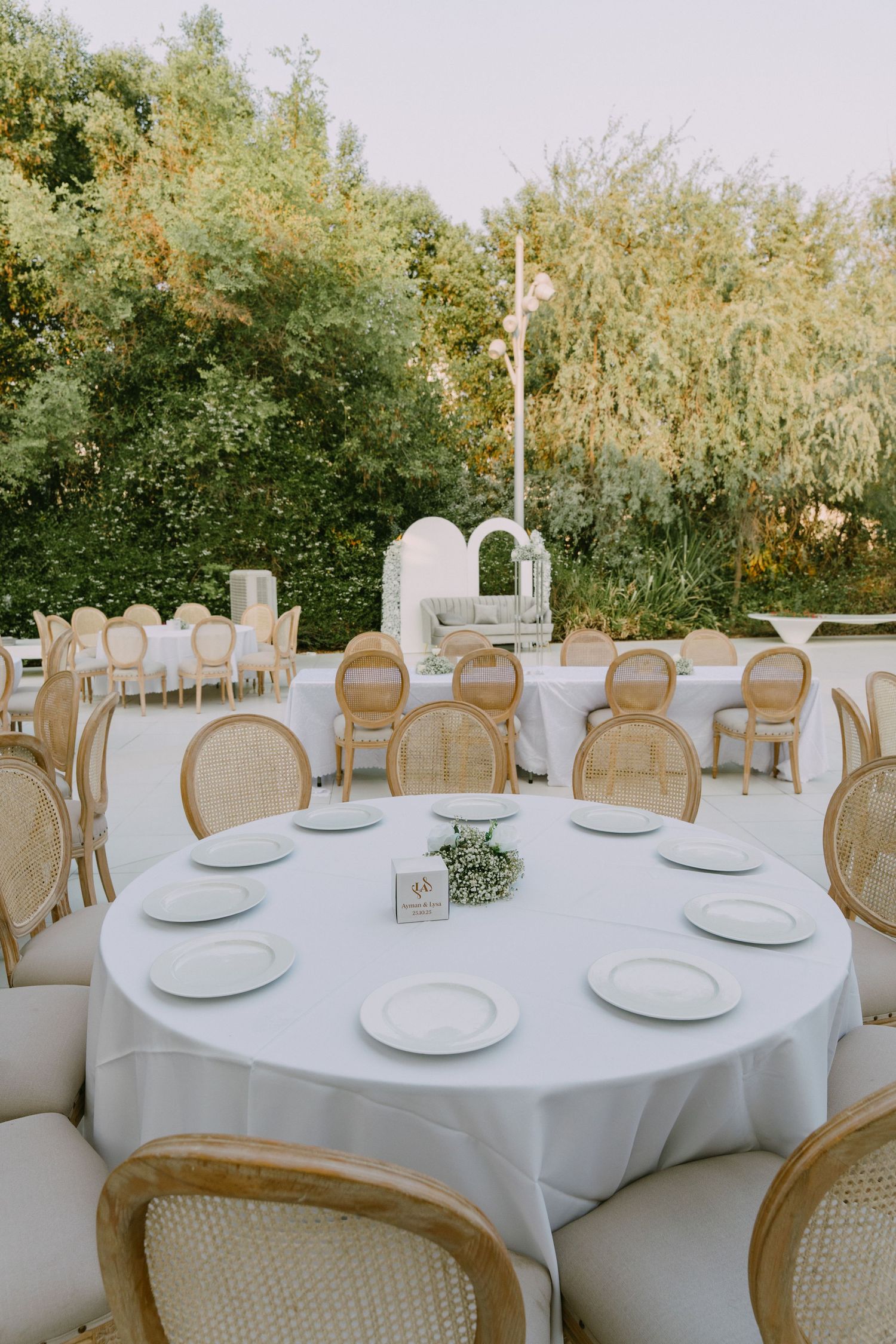 Elegant outdoor wedding reception setup with round tables covered in white linens and French country chairs.
