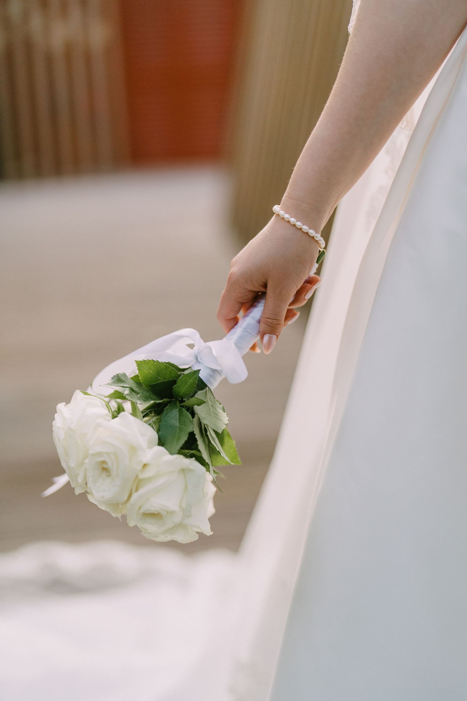 A delicate hand wearing a bracelet holds a small bouquet of white roses against a flowing white dress.