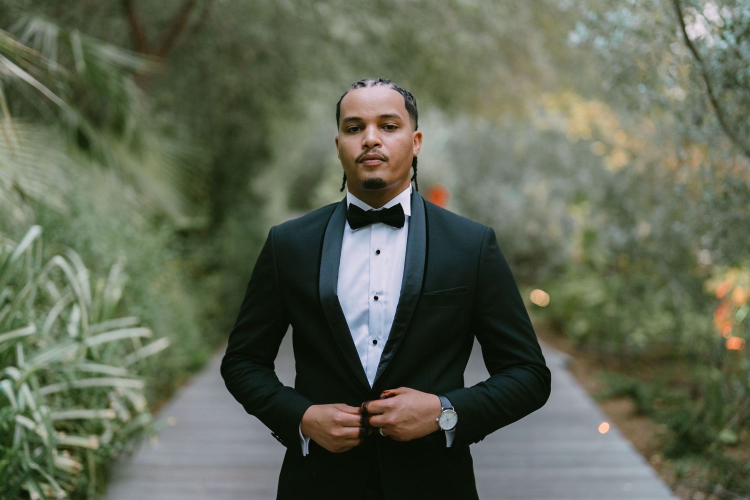A groom in a classic black tuxedo and bow tie poses in a scenic outdoor garden setting during a formal wedding shoot.