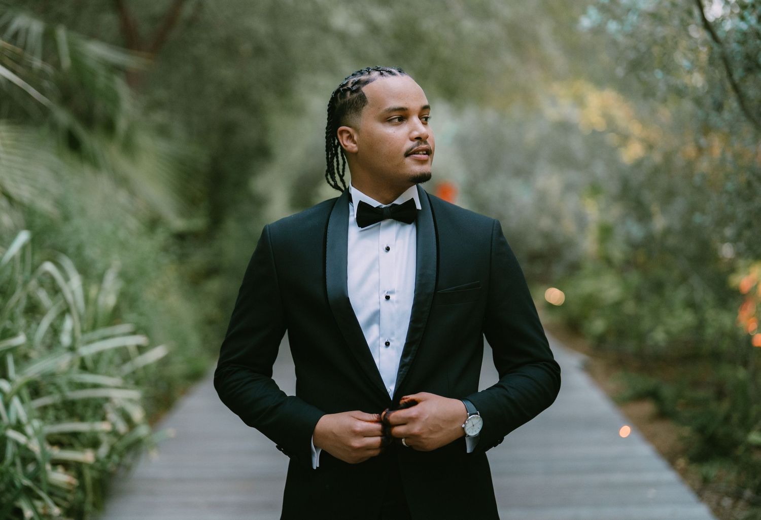 A person in a black tuxedo and bowtie stands on a tree-lined path during an outdoor photoshoot.