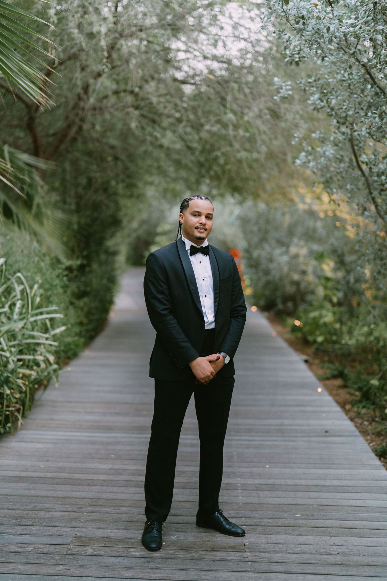 A sequence of formal portraits showing someone in a black tuxedo posing playfully on a wooden boardwalk surrounded by trees.