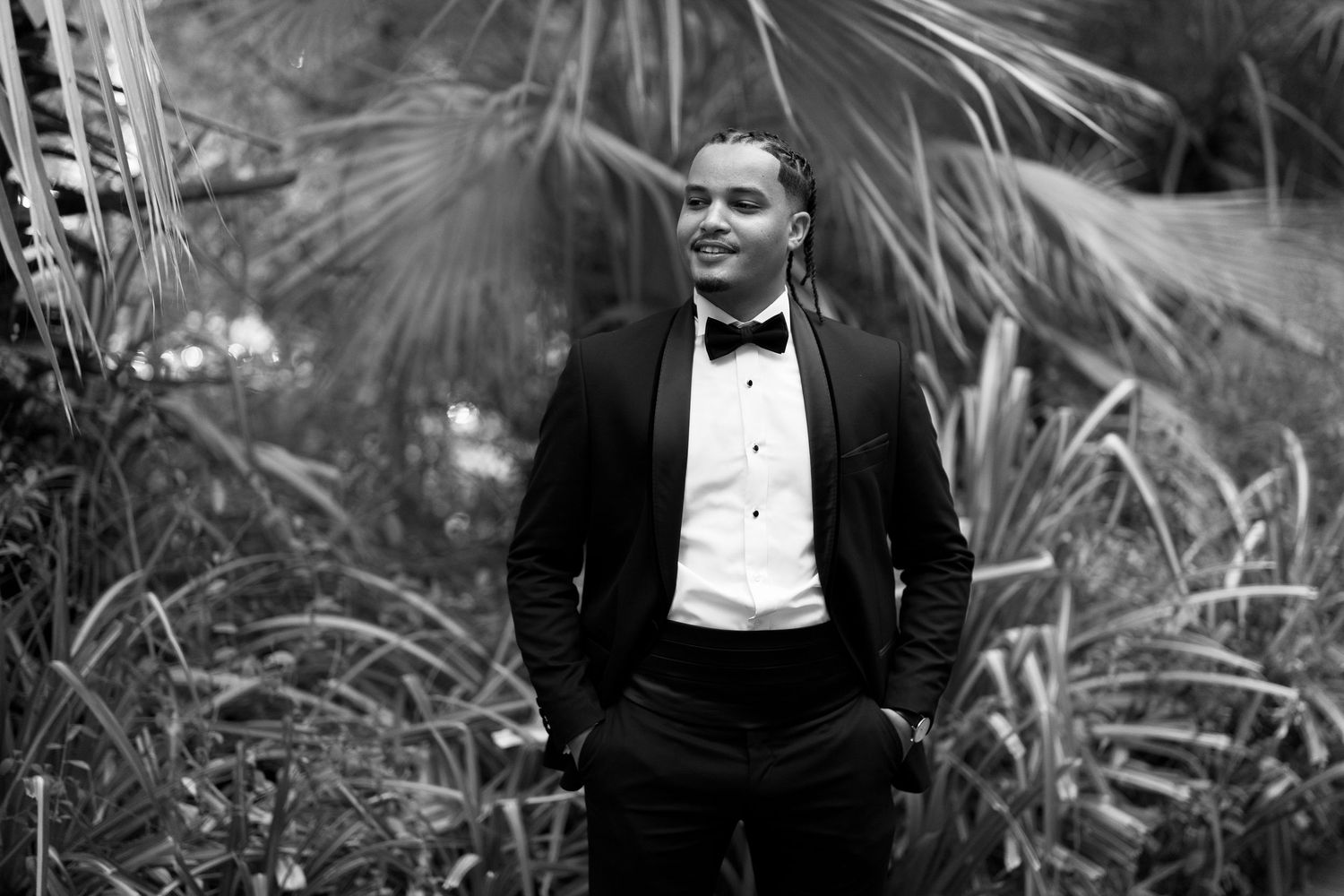 A person in a black tuxedo and bow tie stands confidently in a garden setting with palm fronds in the background.