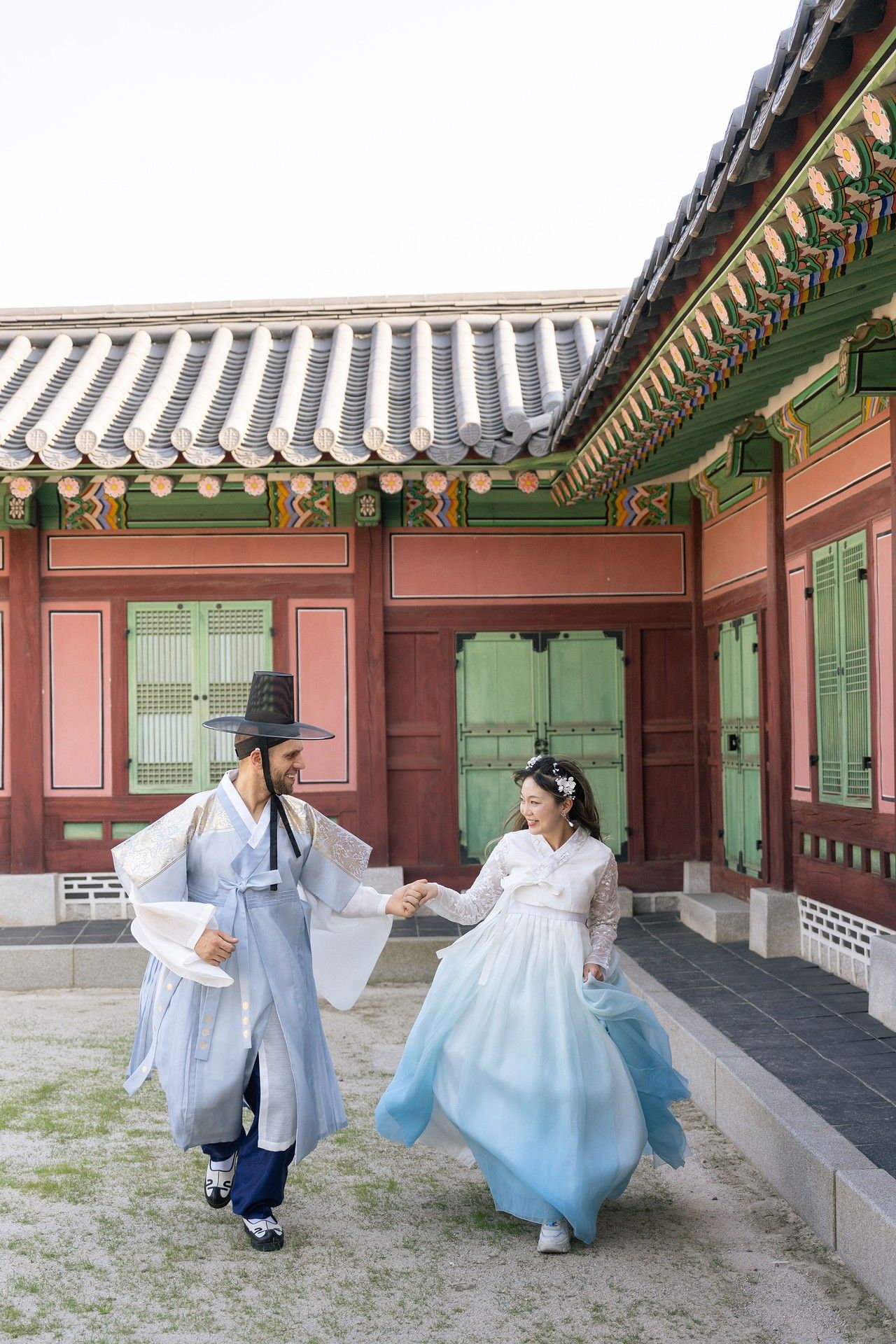 Two people in traditional Korean hanbok outfits dance playfully along a palace corridor with traditional architecture.