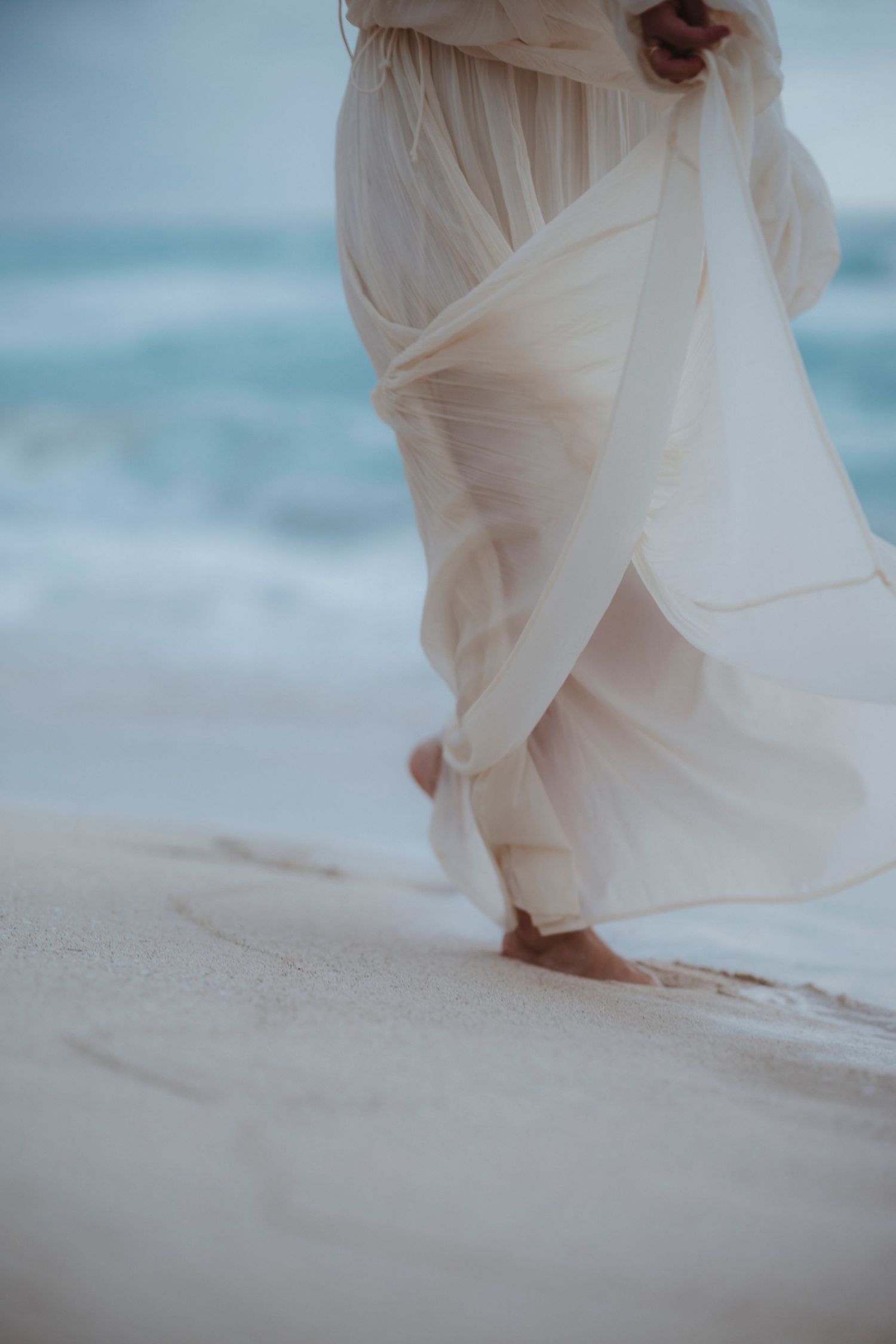 A person in a flowing red dress walks barefoot along a white sandy beach with turquoise ocean waves in the background.