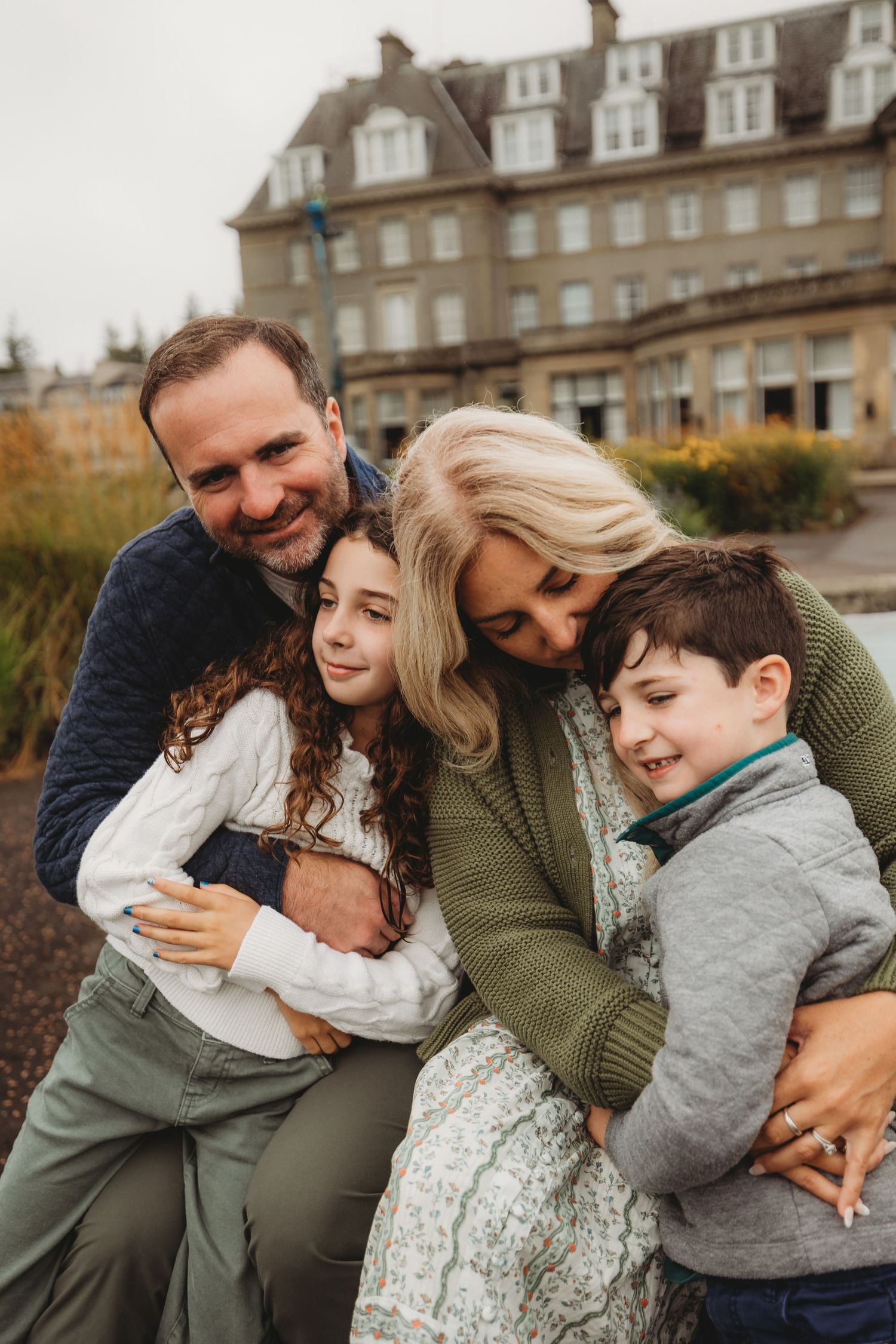 A loving family shares a warm embrace outdoors in front of a historic building on an autumn day.