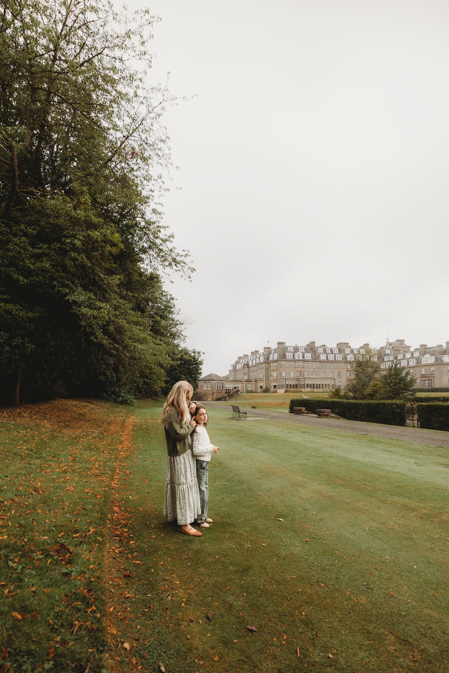 Two figures stand on a manicured lawn near historic buildings and autumn trees on an overcast day.