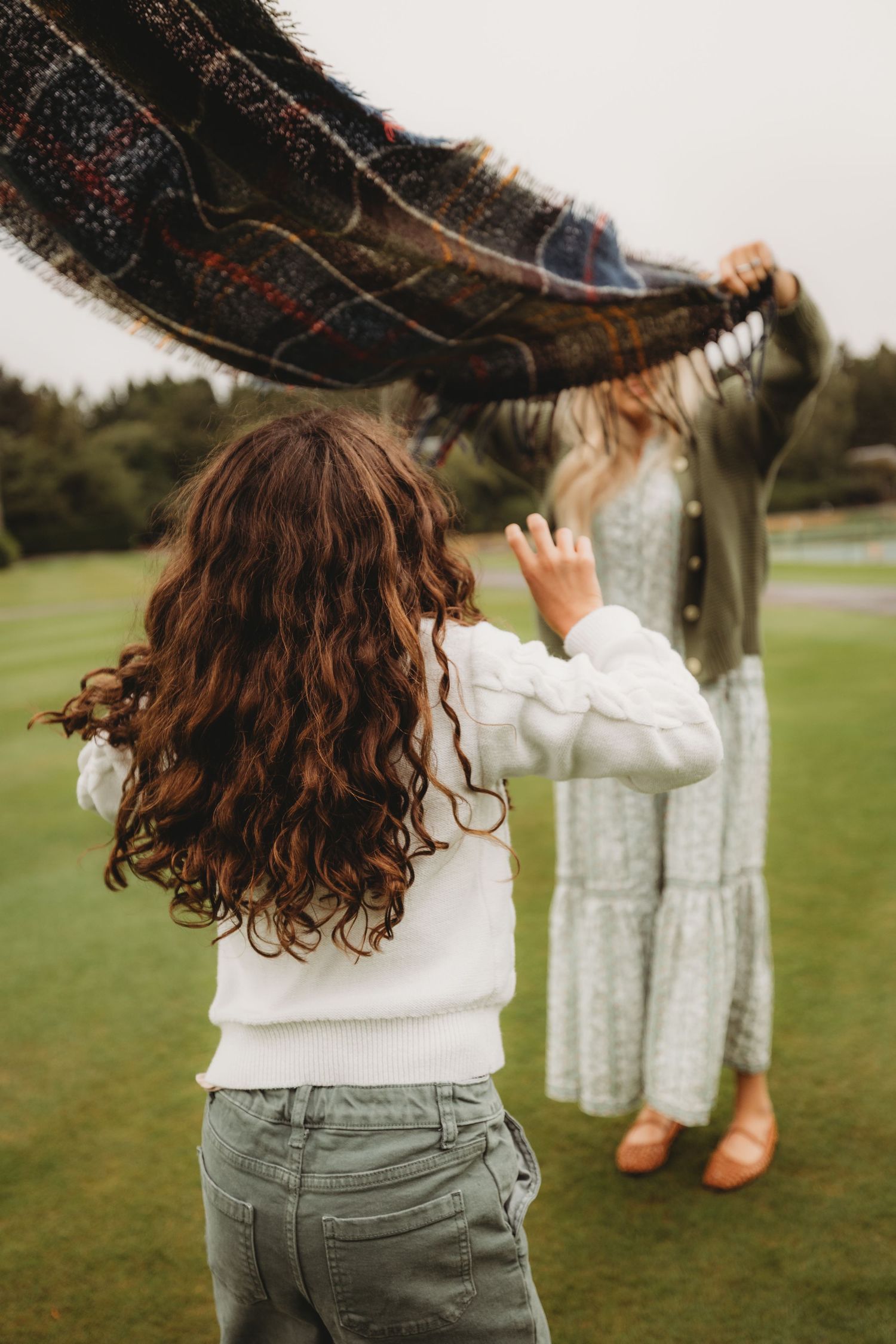A person in a white sweater reaches up towards a dark patterned blanket being held up on a grassy lawn.